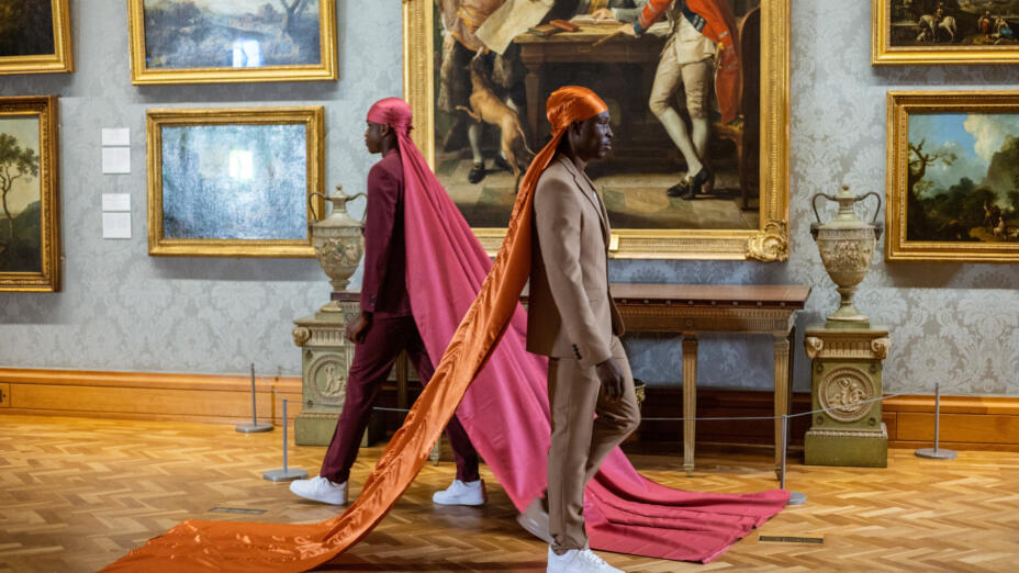 Two young black men are walking past each other. In the background, there are ornate 18th and 19th century paintings in gold frames from the National Museum Wales collection. The young man on the left is dressed in a plum red suit and a similar coloured durag which flows down into floor-length silk. The man on the right is in a tan suit, with a bright orange durag which similarly flows down into floor-length silk