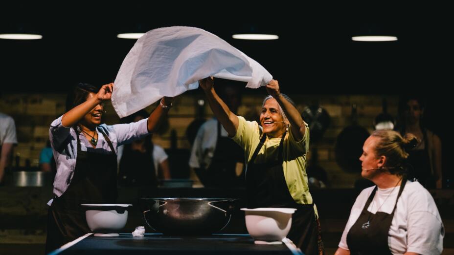 Three women are on a stage, each with headset microphones and wearing aprons. Two of the women are standing up, lifting a white cloth over a table set with mixing bowls.