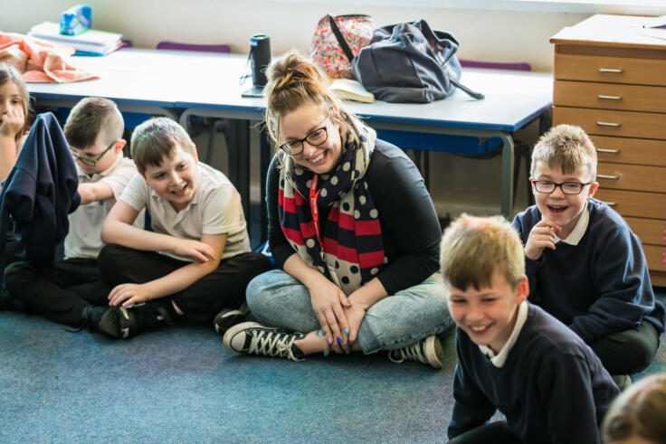 A young woman is sitting cross-legged on the ground, with five primary school aged children sitting either side of her. They are all smiling and looking at one of the children