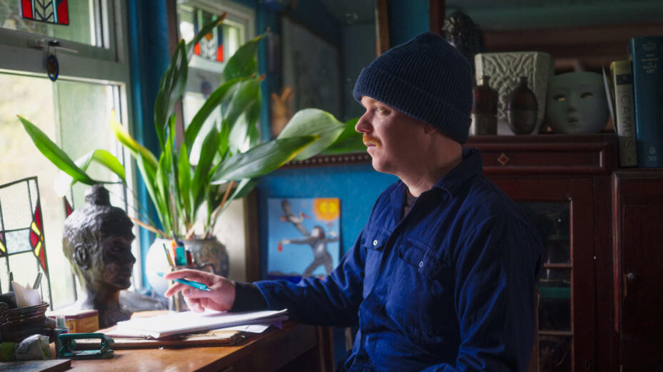 Artist wears blue jumpsuit and beanie while working at a desk surrounded by books and looking out a stained glass window