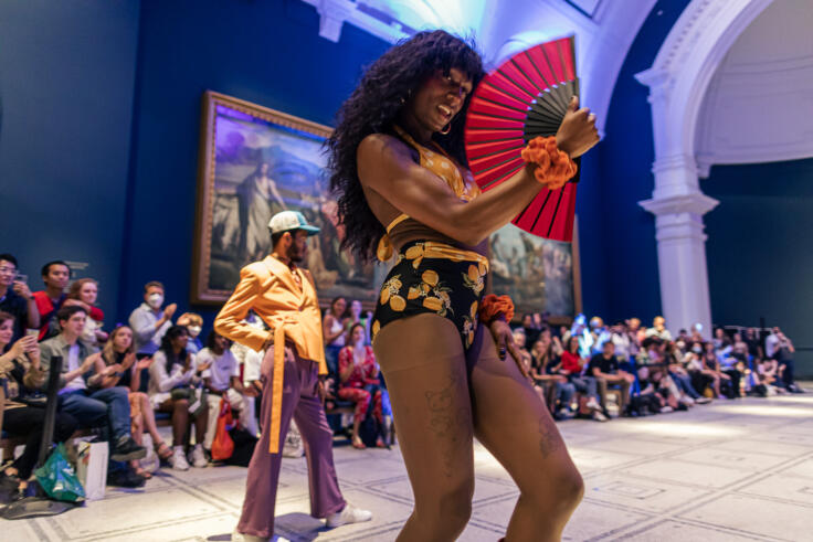 Two young performers are standing in a dark blue, ornate room, with a busy seated crowd in the background. The performer at the front is wearing bright yellow and black clothes and is holding a red fan and looks to be mid-dance. The performer at the back has a hand on their hip, and is wearing a bright yellow jacket with a long belt, purple trousers and a baseball cap