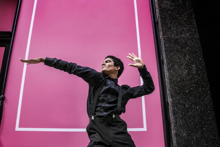 A young man all dressed in black is posing against a bright pink wall. He is in a shirt with suspenders and his right arm is stretched out in front of him with his left reaching backwards behind him