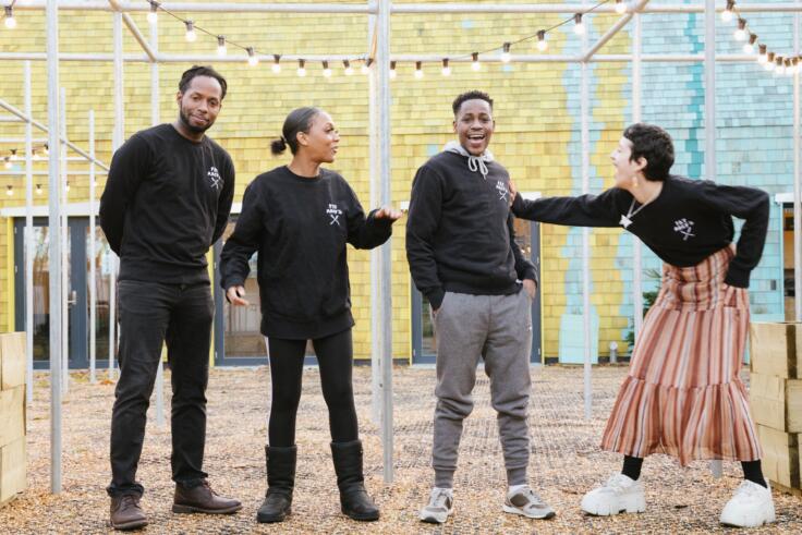 Four young people are standing and posing for a photo. One person is laughing and being playfully pushed by another