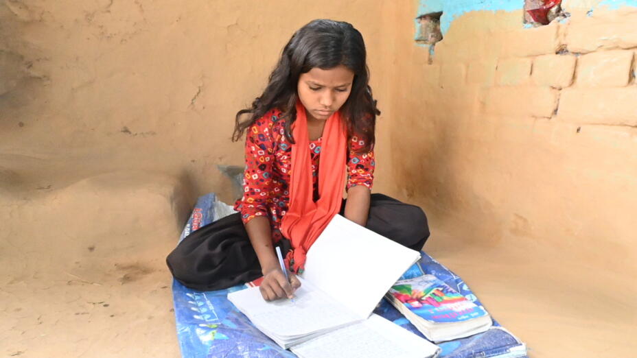 Young girl with a red shawl sits outside on a blue rug reading and doing school work.
