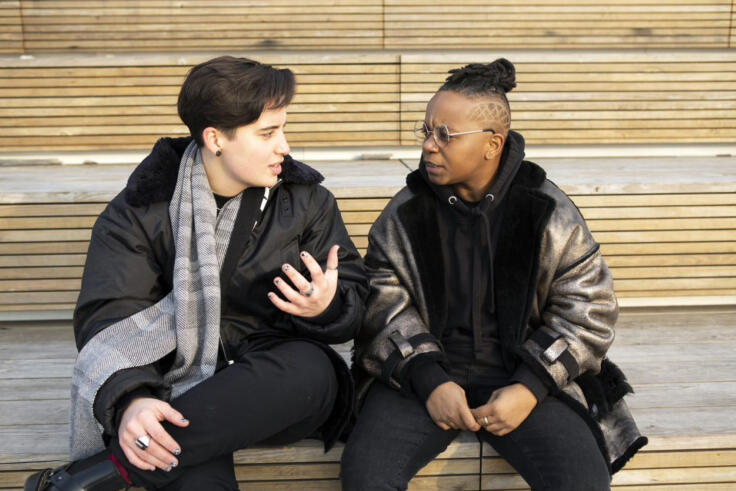 Two people are sat on a wooden structure outside, in conversation