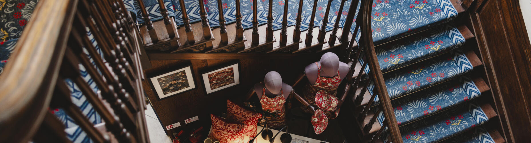Staircase covered in William Morris dark blue floral print, shot from above. At the bottom of the stairs is a table with William Morris prints and items for sale.