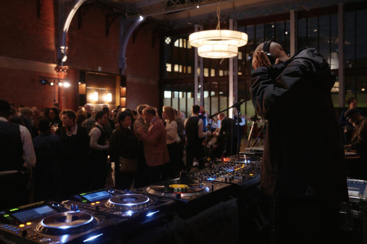 A DJ is holding his headphones, in front of a standing crowd at a reception event.