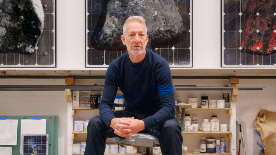 Artist sits on a stool in his studio in front of three works featuring triangular stones in grey, black and red colour palette