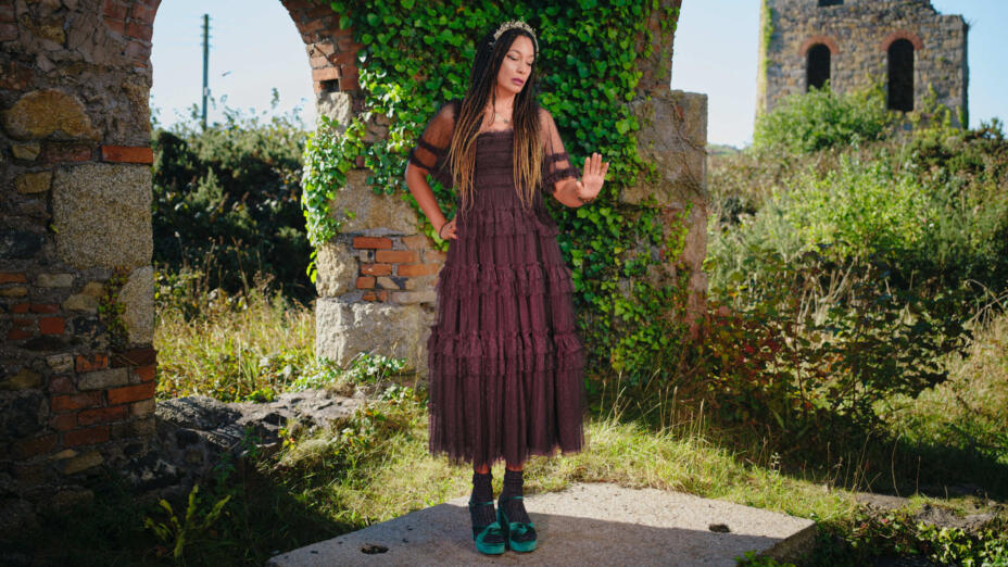 Composer stands in a diaphanous burgundy dress and turquoise sandals amidst ruins of stone, medieval buildings