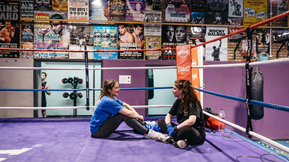Two girls sit on the floor of a boxing ring chatting together. The walls of the gym are covered in posters.