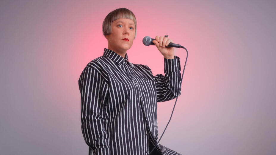 Artist wears a black and white striped, long-sleeved button down shirt. She holds a microphone in one hand and stands in front of a white wall. A sift pick light glows behind her