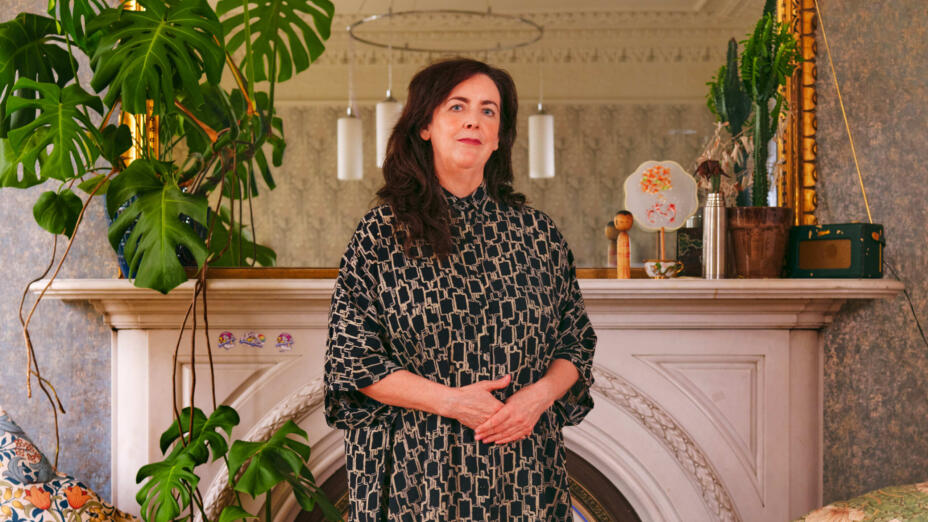 Artist stands before a white fireplace in front of a gold framed mirror and surrounded by lush green plants