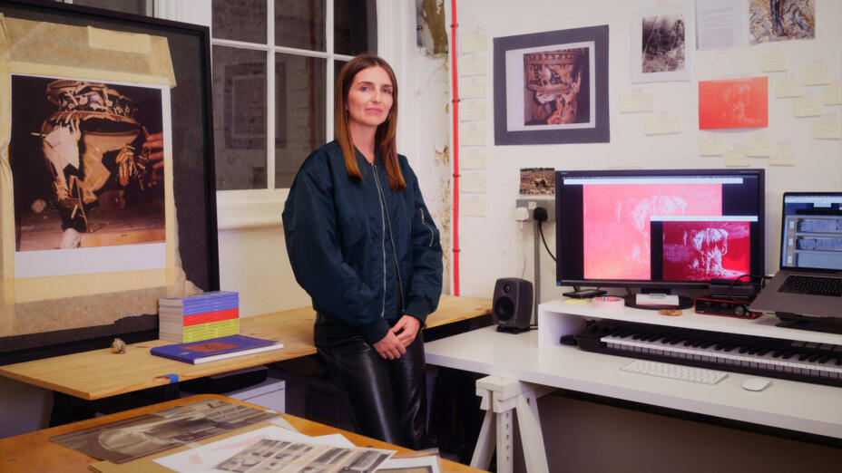 Artist stands at a wooden desk in a studio with white-painted brick walls surrounded by images and posters related to her works