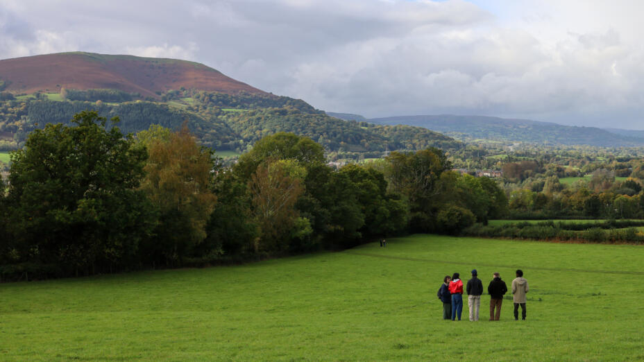 A scenic shot of Castle Meadows in Wales, showing a large green field with five people standing in the distance. In the background is a cloudy blue sky, a thick forest of trees and large red hills.